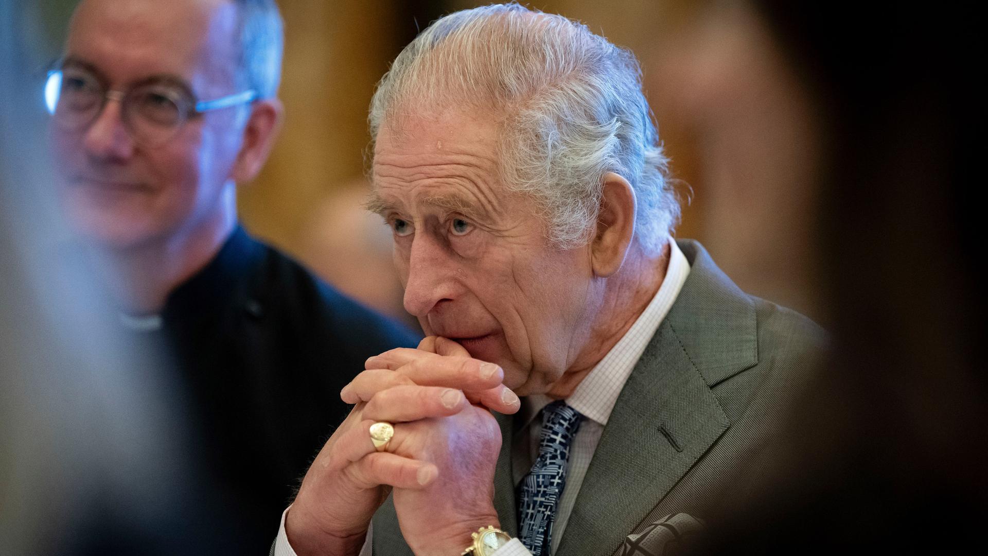 Der britische König Charles III. sitzt nachdenklich am Tisch. Die Hände hat er vor seinem Kinn gefaltet. 