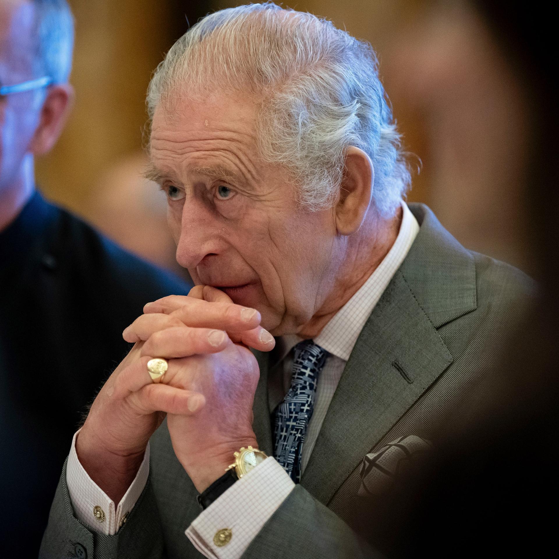 Der britische König Charles III. sitzt nachdenklich am Tisch. Die Hände hat er vor seinem Kinn gefaltet. 
