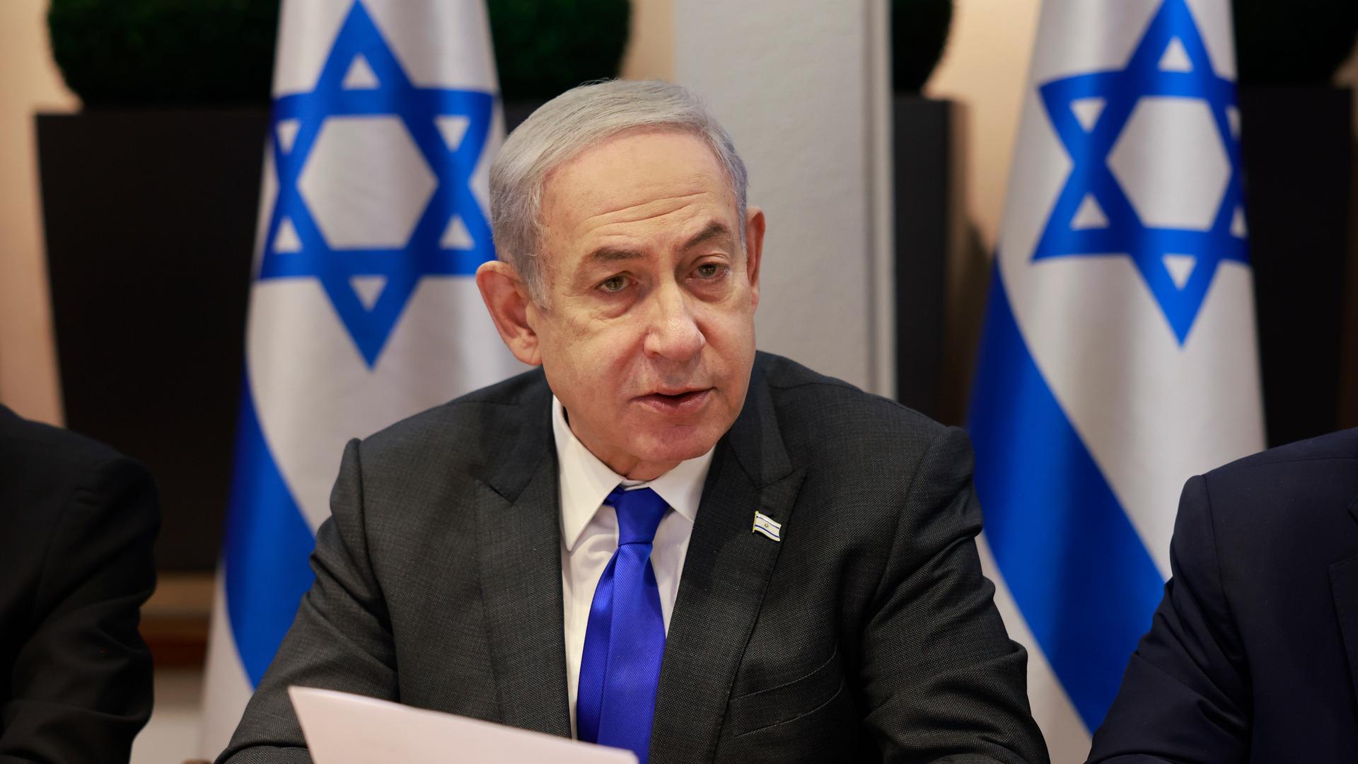 Israels Ministerpräsident Benjamin Netanyahu sitzt mit einem Zettel in der Hand vor Israel-Fahnen