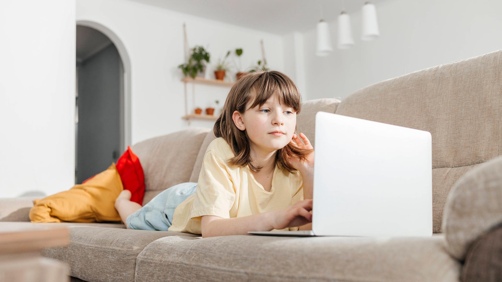 Ein junges Mädchen liegt auf einer Couch und schaut auf einen Laptop.