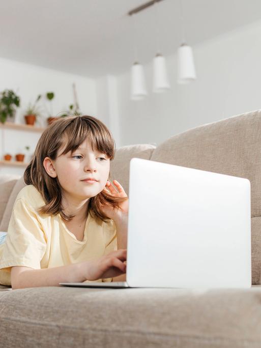 Ein junges Mädchen liegt auf einer Couch und schaut auf einen Laptop.