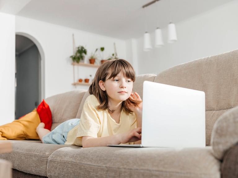 Ein junges Mädchen liegt auf einer Couch und schaut auf einen Laptop.