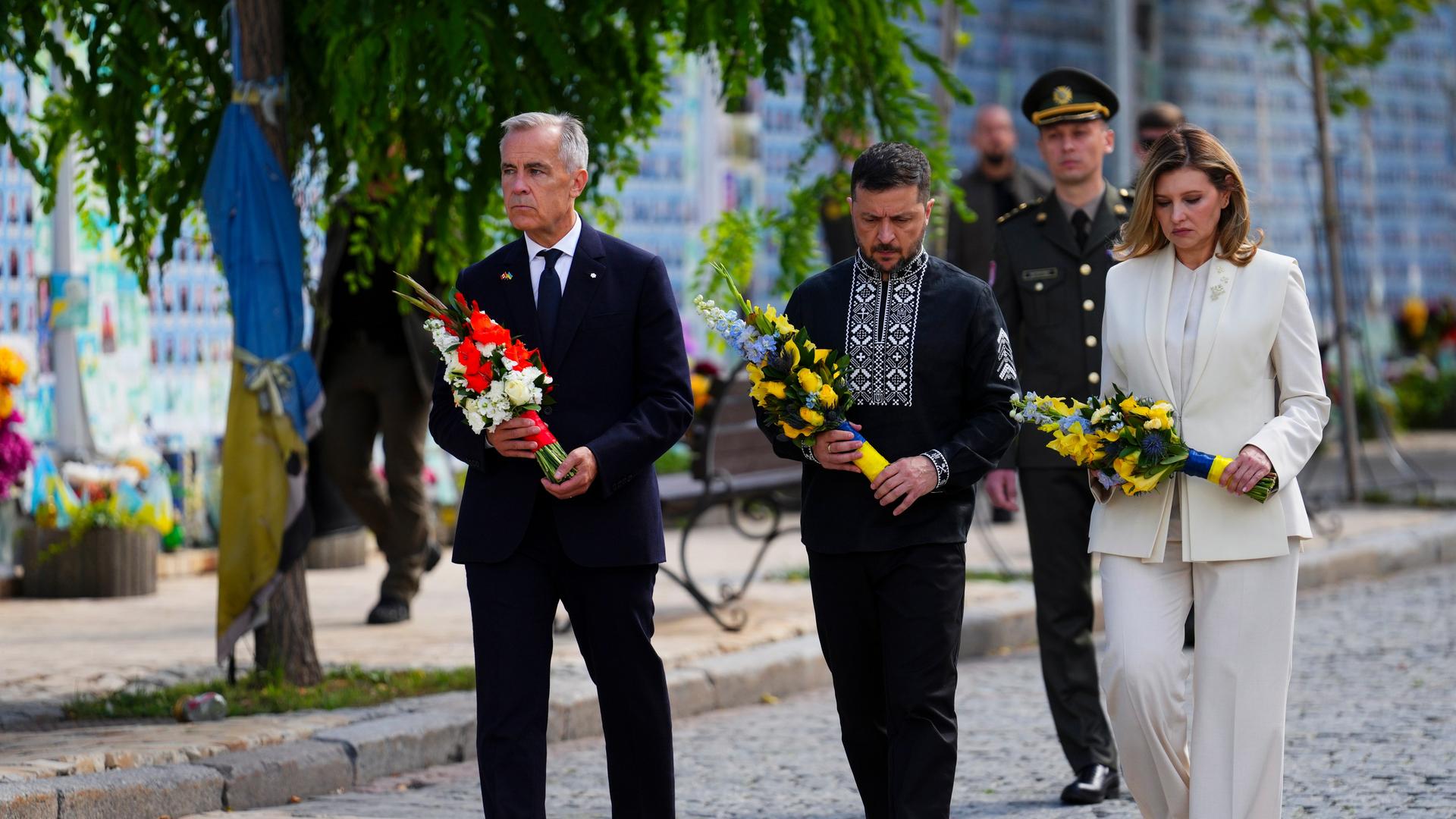 Kanadas Premierminister Carney, der ukrainische Präsident Selenskyj und seine Ehefrau Olena gehen nebeneinander her. Sie schauen ernst und tragen Blumen in den Händen. 