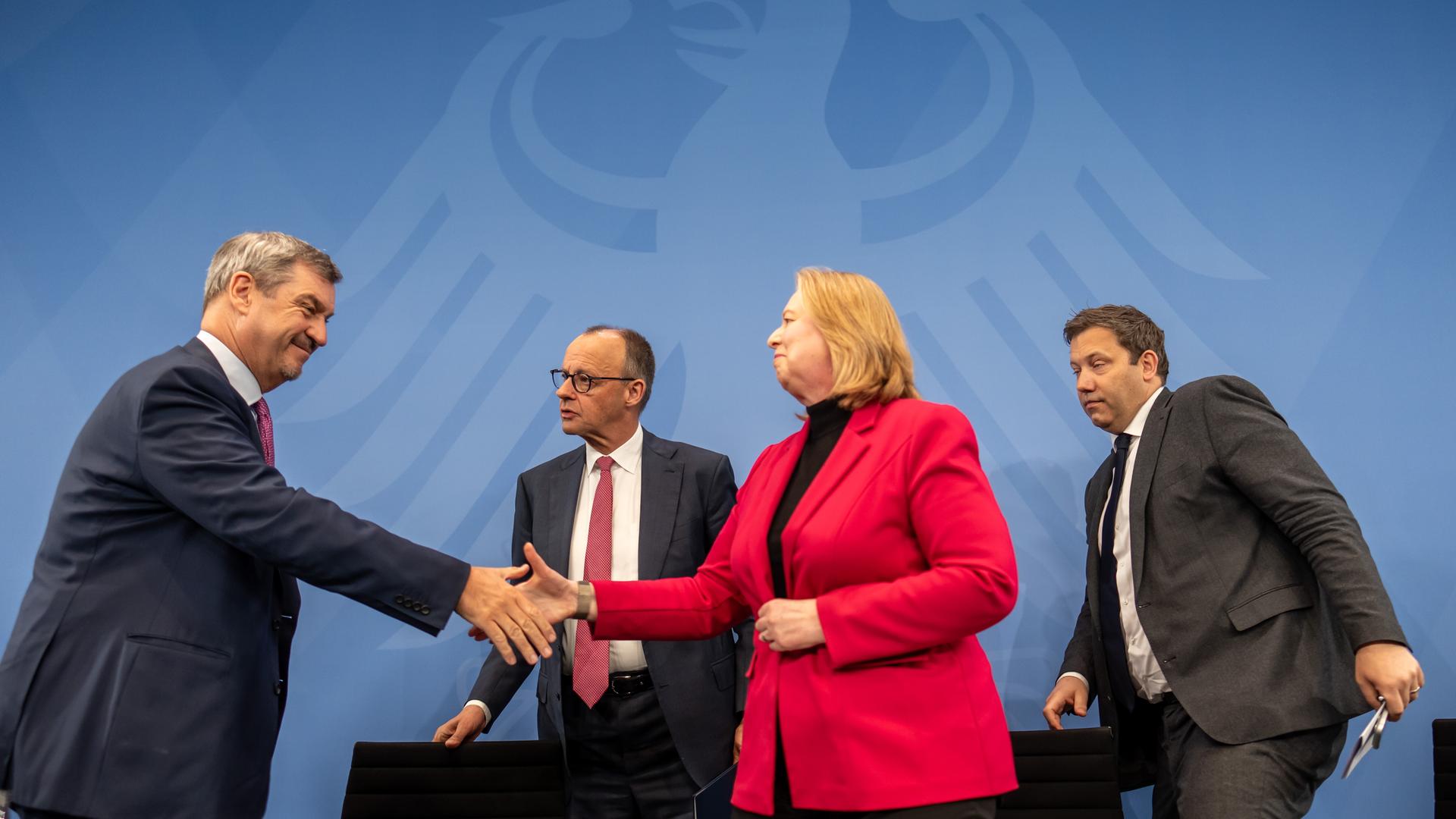 CSU-Chef Söder und die SPD-Vorsitzende Bas schütteln sich die Hände. Dahinter stehen Kanzler Merz und SPD-Co-Chef Klingbeil. Im Hintergrund ist der Bundesadler auf blauem Grund zu sehen. 
