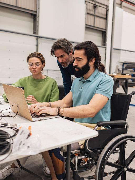 Ein bärtiger Mann sitzt im Rollstuhl am Schreibtisch und tippt auf der Tastatur eines Laptops, daneben liegen Kabel, im Hintergrund ist eine Werkhalle zu sehen. Ein Mann und eine Frau blicken ihm über die Schulter.