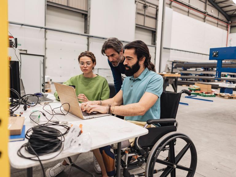 Ein bärtiger Mann sitzt im Rollstuhl am Schreibtisch und tippt auf der Tastatur eines Laptops, daneben liegen Kabel, im Hintergrund ist eine Werkhalle zu sehen. Ein Mann und eine Frau blicken ihm über die Schulter.