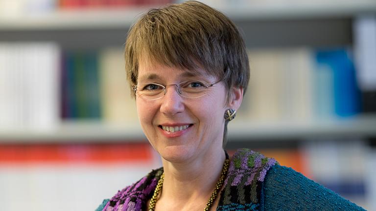 Die Soziologin Nicole Mayer-Ahuja, mit Brille, kurzem Haar, in einer blauen Strickbluse sitzt vor einem Bücherregal und lächelt freundlich in die Kamera.