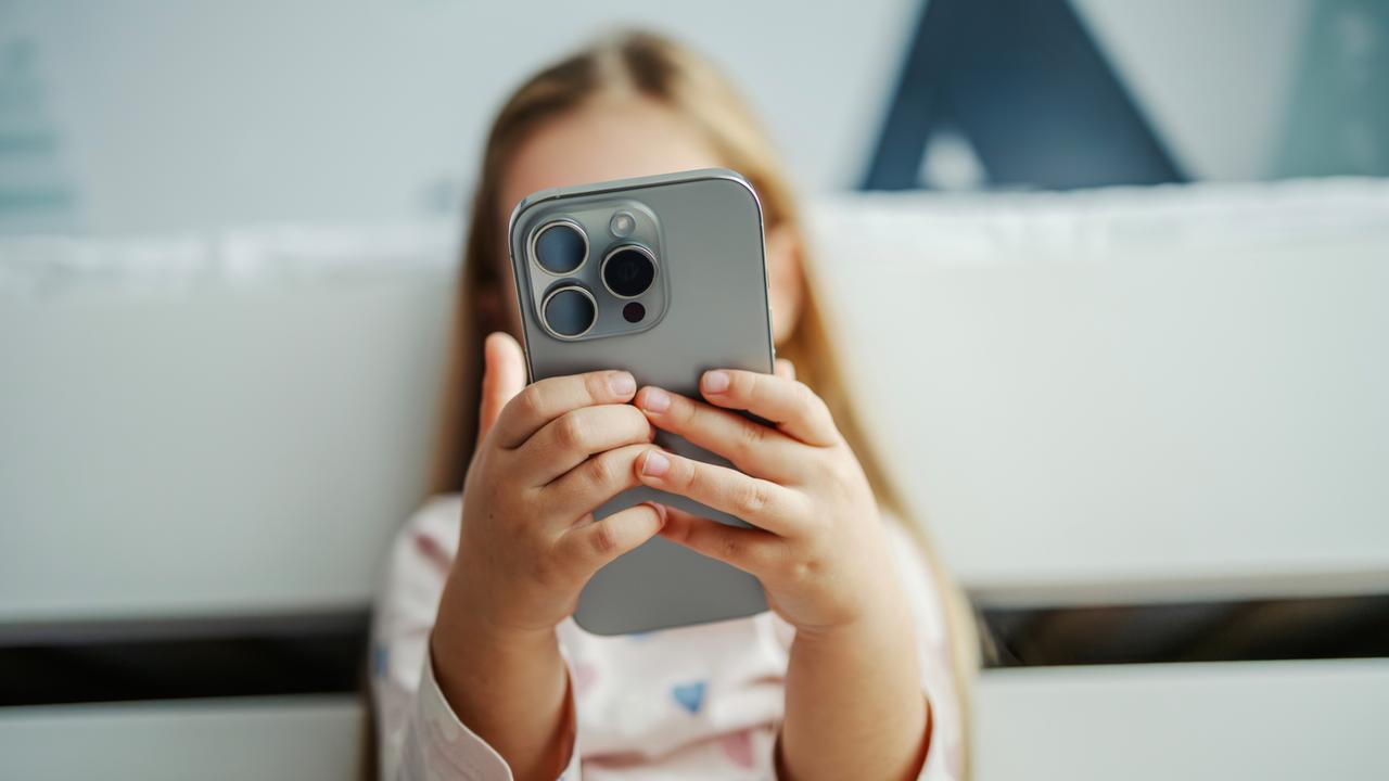 Ein Kind hält ein modernes graues Smartphone in der Hand. Das Smartphone verdeckt das Gesicht des Kindes. Das Kind im Hintergrund ist nur unscharf zu erkennen.