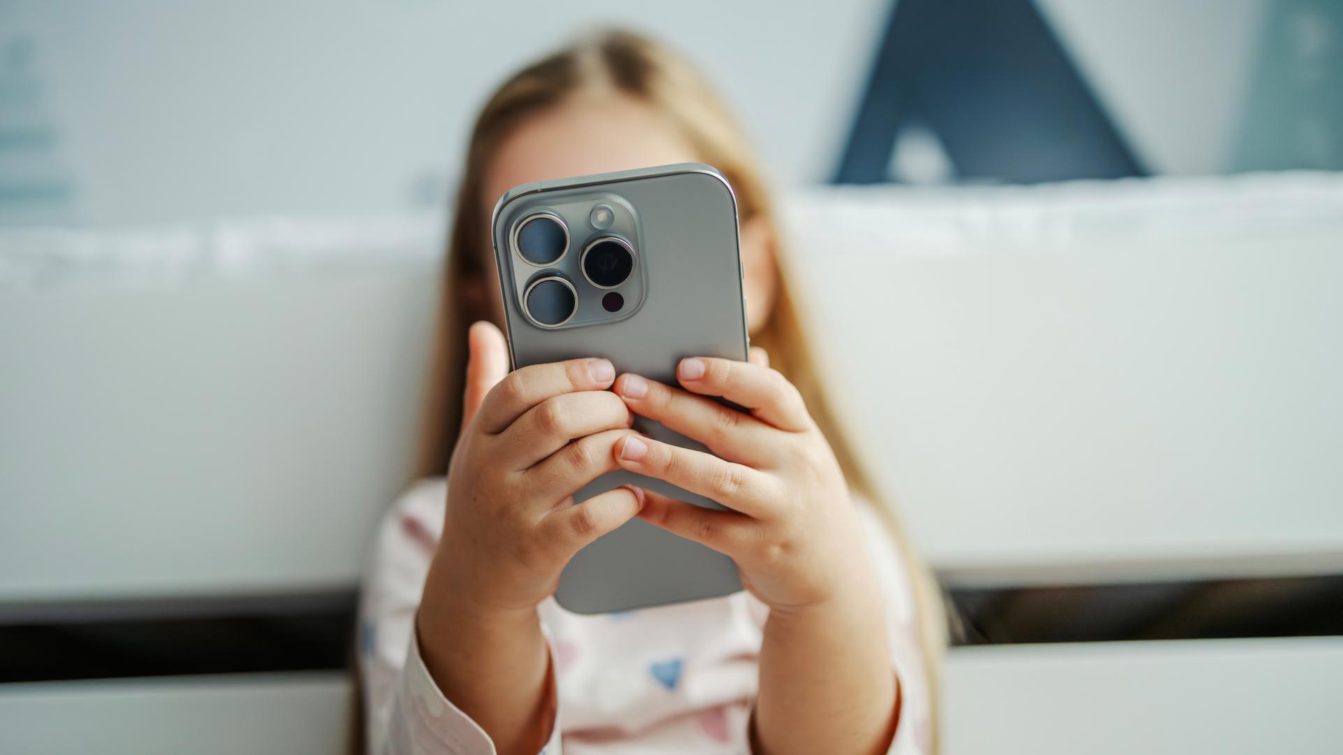 Ein Kind hält ein modernes graues Smartphone in der Hand. Das Smartphone verdeckt das Gesicht des Kindes. Das Kind im Hintergrund ist nur unscharf zu erkennen.