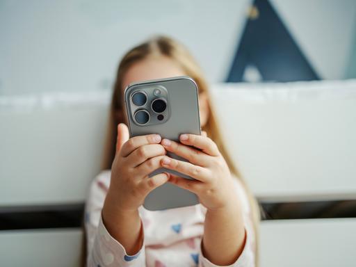 Ein Kind hält ein modernes graues Smartphone in der Hand. Das Smartphone verdeckt das Gesicht des Kindes. Das Kind im Hintergrund ist nur unscharf zu erkennen.