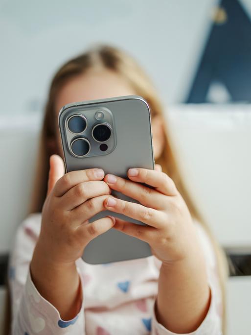 Ein Kind hält ein modernes graues Smartphone in der Hand. Das Smartphone verdeckt das Gesicht des Kindes. Das Kind im Hintergrund ist nur unscharf zu erkennen.