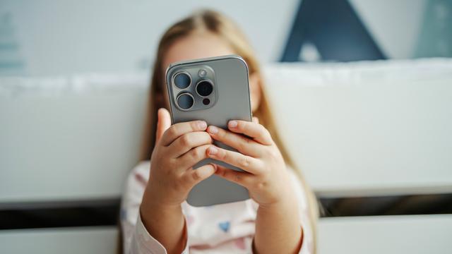 Ein Kind hält ein modernes graues Smartphone in der Hand. Das Smartphone verdeckt das Gesicht des Kindes. Das Kind im Hintergrund ist nur unscharf zu erkennen.