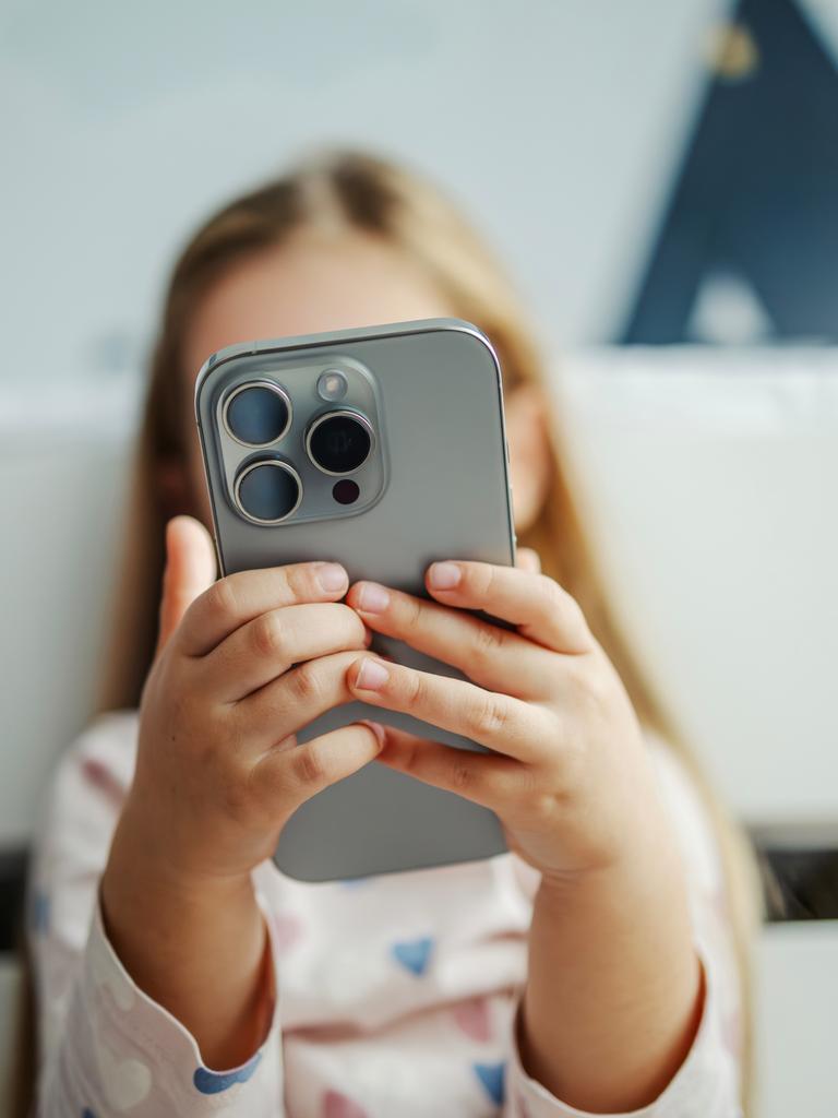 Ein Kind hält ein modernes graues Smartphone in der Hand. Das Smartphone verdeckt das Gesicht des Kindes. Das Kind im Hintergrund ist nur unscharf zu erkennen.