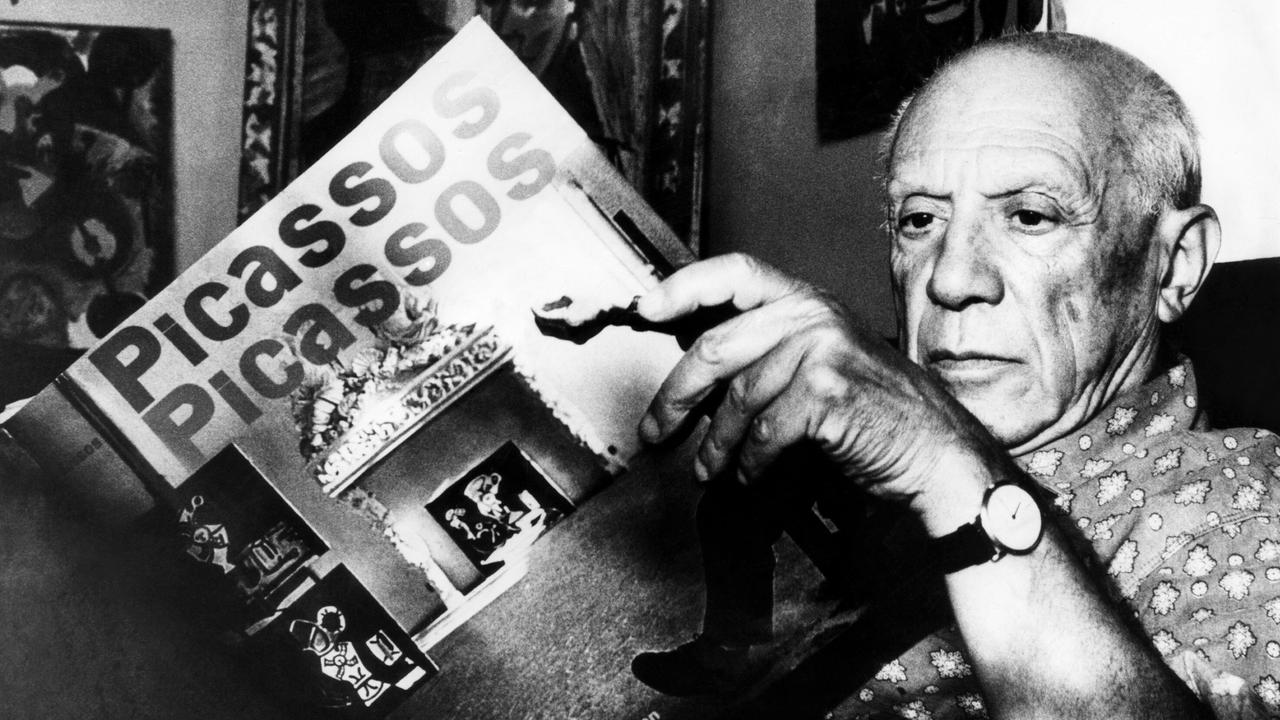 Pablo Picasso sitzt in einem Sessel und hält ein Buch mit der Aufschrift "Picassos" in den Händen.