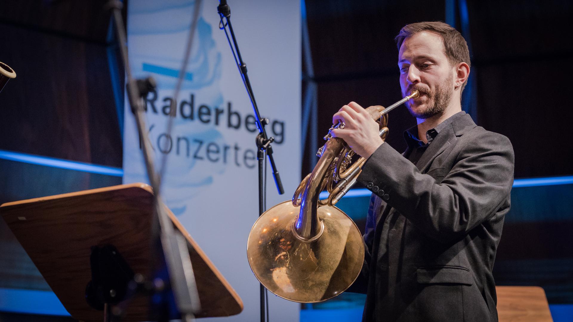 Ein Hornist spielt in einem holzvertäfelten Saal, der bläulich angestrahlt wird. Im Hintergrund hängt ein großes Plakat mit der Aufschrift "Raderberg Konzerte - Deutschlandfunk".