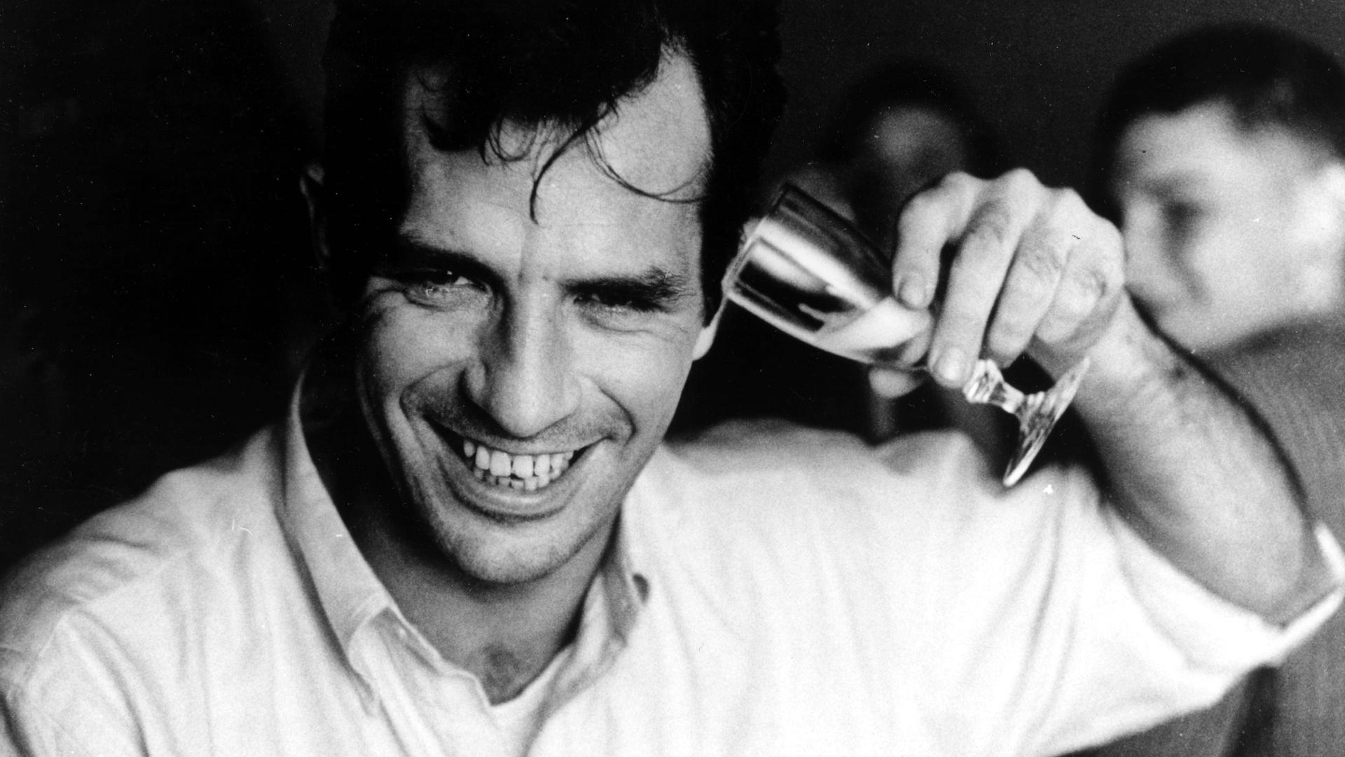 Porträt von Jack Kerouac, der sich lachend ein Sektglas ans Ohr hält.