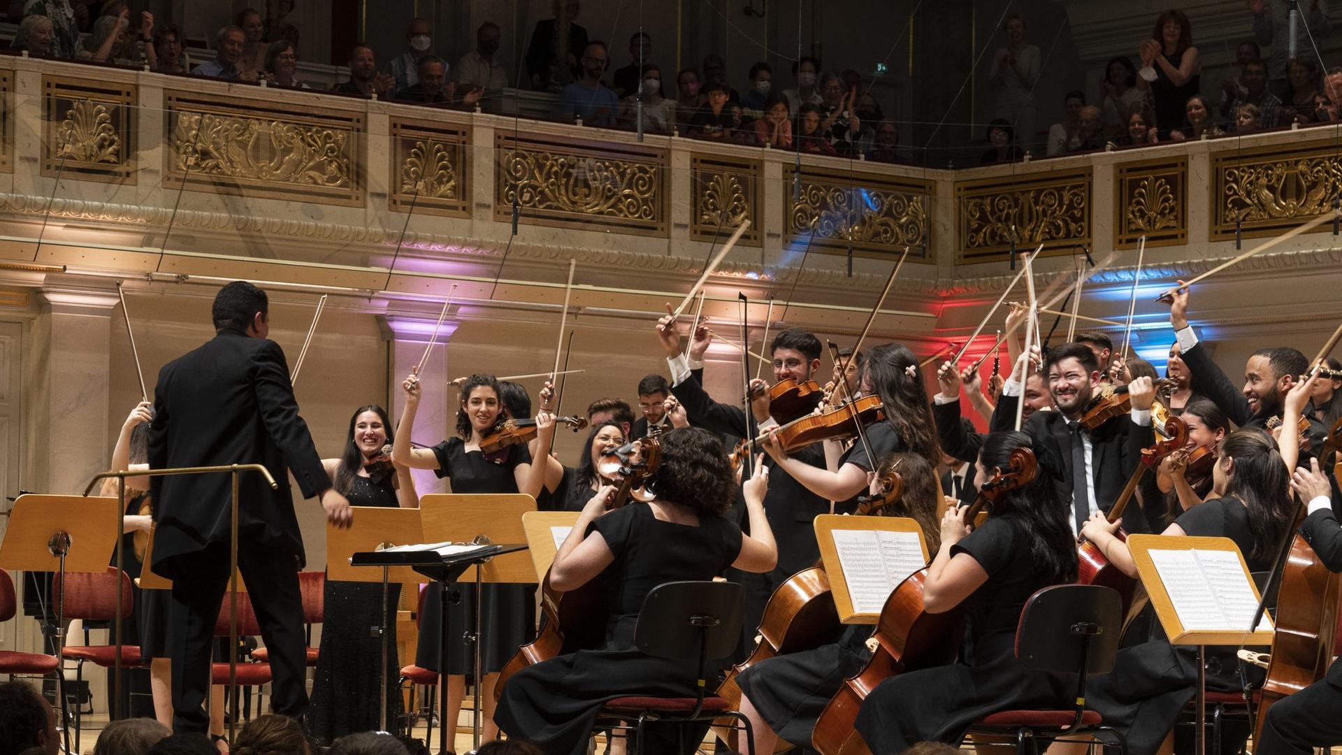 Junger Musikerinnen und Musiker eines Jugendorchesters lassen zum Abschluss eines Stückes die Bögen in die Höhe gestreckt und lachen dem Dirigenten entgegen. Junger Musikerinnen und Musiker eines Jugendorchesters lassen zum Abschluss eines Stückes die Bögen in die Höhe gestreckt und lachen dem Dirigenten entgegen.