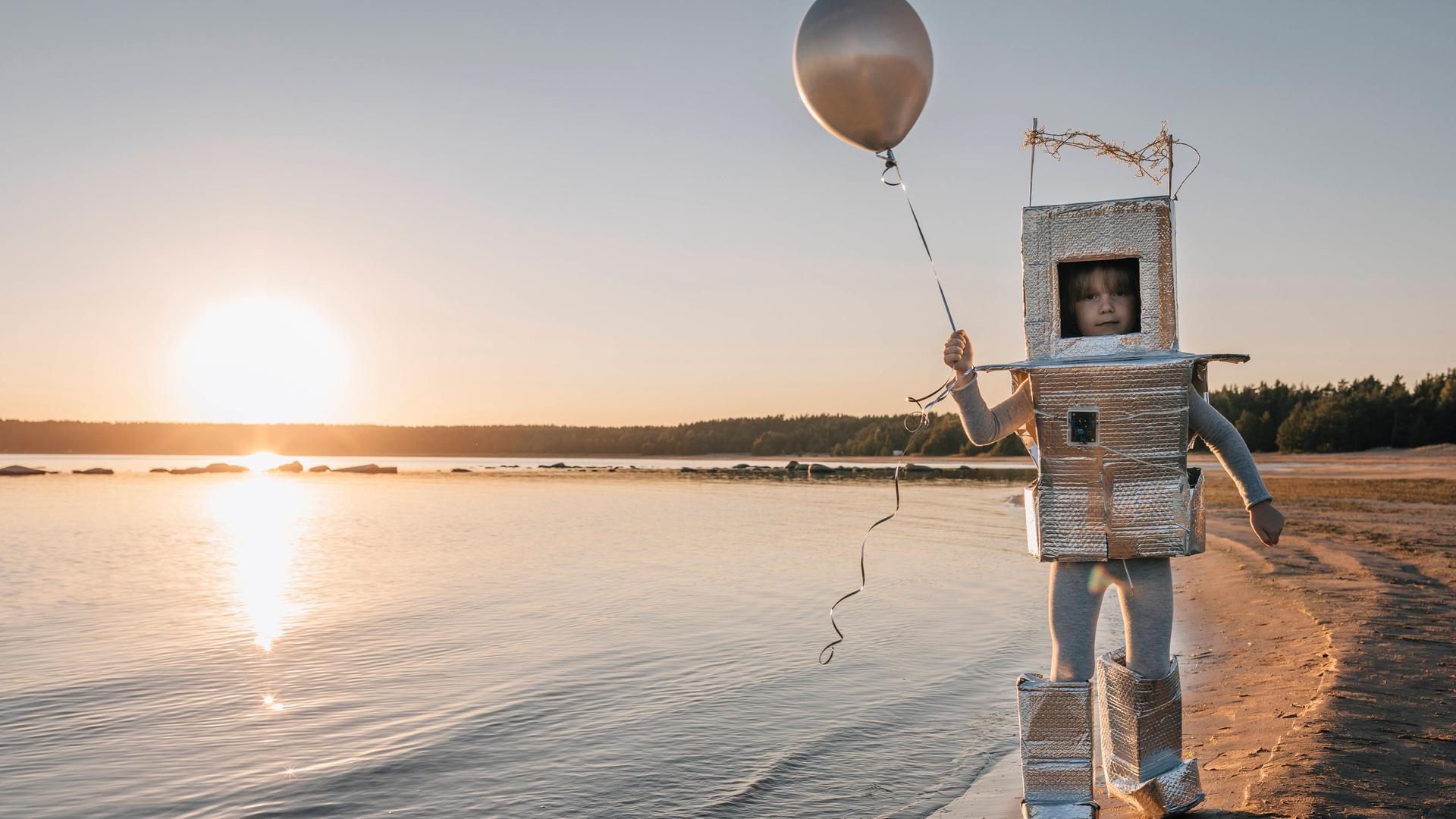 Ein Kind in einem gebastelten Kosmonautenanzug hält einen Ballon in der Hand und läuft am Ufer eines Sees entlang.