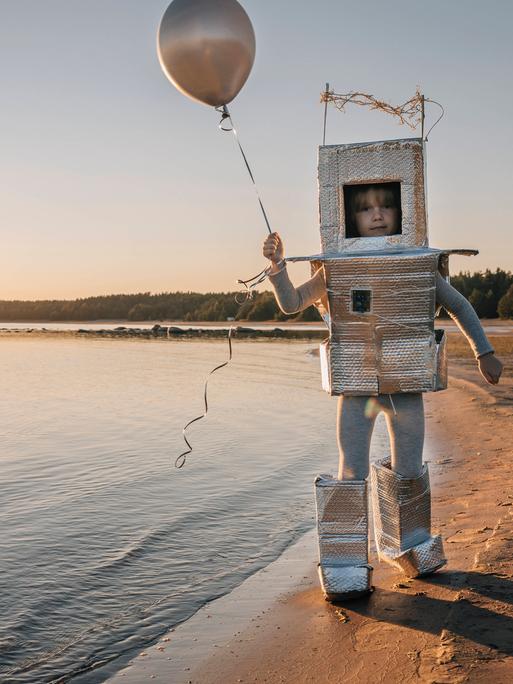 Ein Kind in einem gebastelten Kosmonautenanzug hält einen Ballon in der Hand und läuft am Ufer eines Sees entlang.