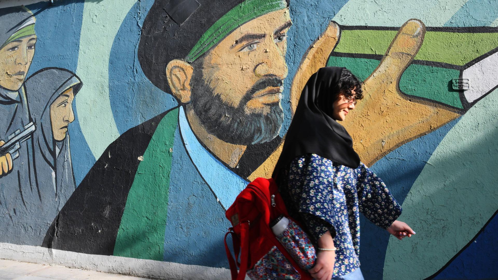 Eine junge Frau im Koptuch und dunklen Haaren die herausschauen geht eine Straße in Teheran entlang, vorbei an einem politischen Graffiti an einer Wand, Iran 2024.