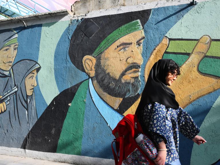 Eine junge Frau im Koptuch und dunklen Haaren die herausschauen geht eine Straße in Teheran entlang, vorbei an einem politischen Graffiti an einer Wand, Iran 2024.