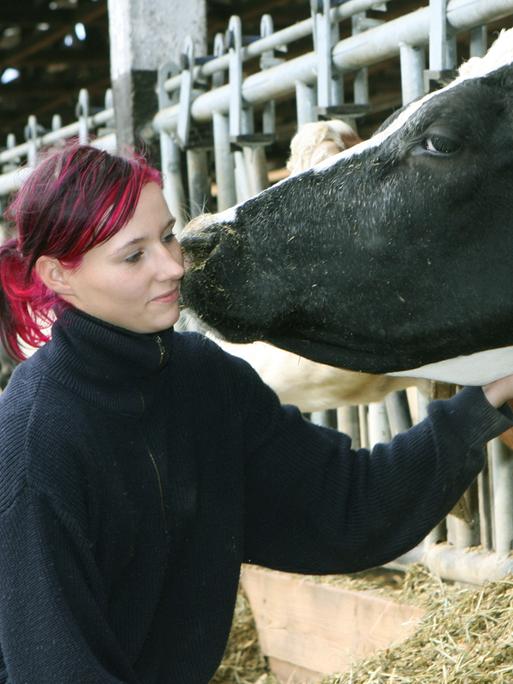 Eine junge Frau mit violett getönten Haaren streichelt eine Kuh, die ihren Kopf aus dem Gatter des Stalls streckt.