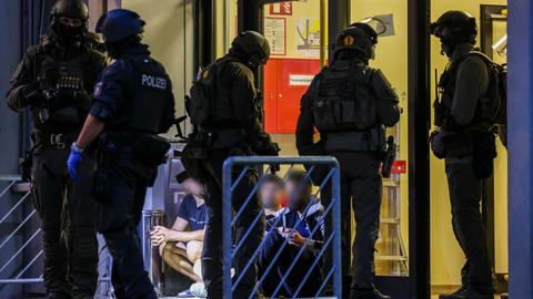 Es ist dunkel. Mehrere Polizisten in Kampfausrüstung stehen am Eingang des Gebäudes. In einem Lichkegel sitzen drei Flüchtlinge, deren Gesicher verpixelt sind. 
