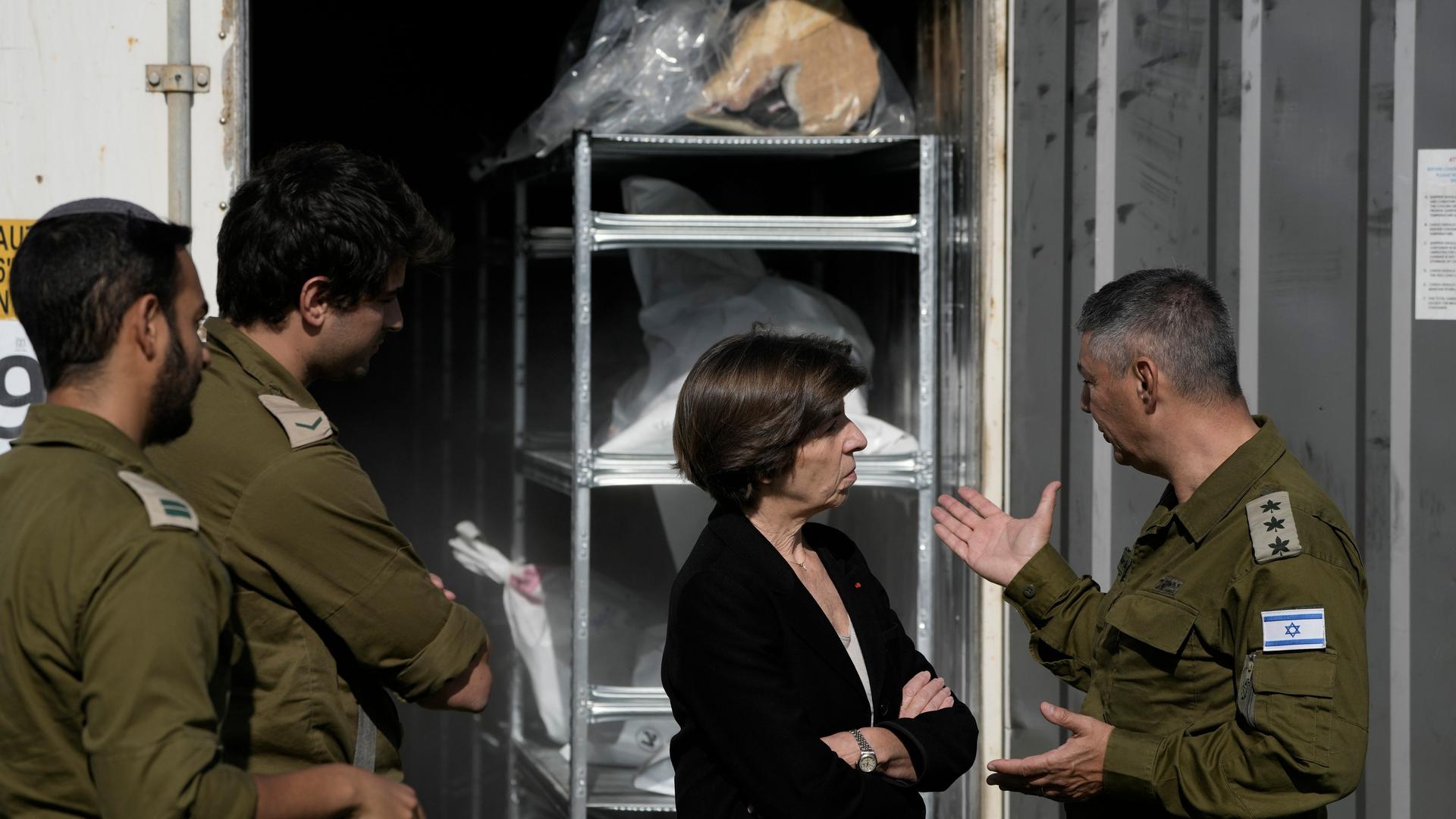 Die französische Außenministerin Catherine Colonna unterhält sich mit einem Offizier. Hinter ihr sind Container zu sehen, in denen die Leichen von Opfern des Terrorangriffs der Hamas aufbewahrt werden.