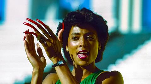 Frau mit kurzen dunklen Haaren in Sportoutfit klatscht in die Hände. An der linken Hand trägt sie extrem lange, rot lackierte Fingernägel