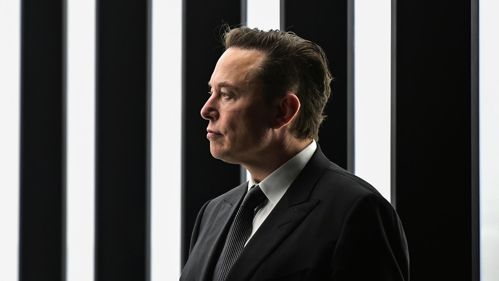 Elon Musk steht vor einem schwarz weiß gestreiften beleuchteten Hintergrund, trägt einen schwarzen Anzug und schaut zur Seite