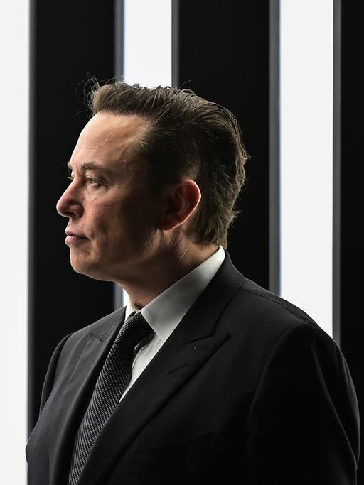 Elon Musk steht vor einem schwarz weiß gestreiften beleuchteten Hintergrund, trägt einen schwarzen Anzug und schaut zur Seite