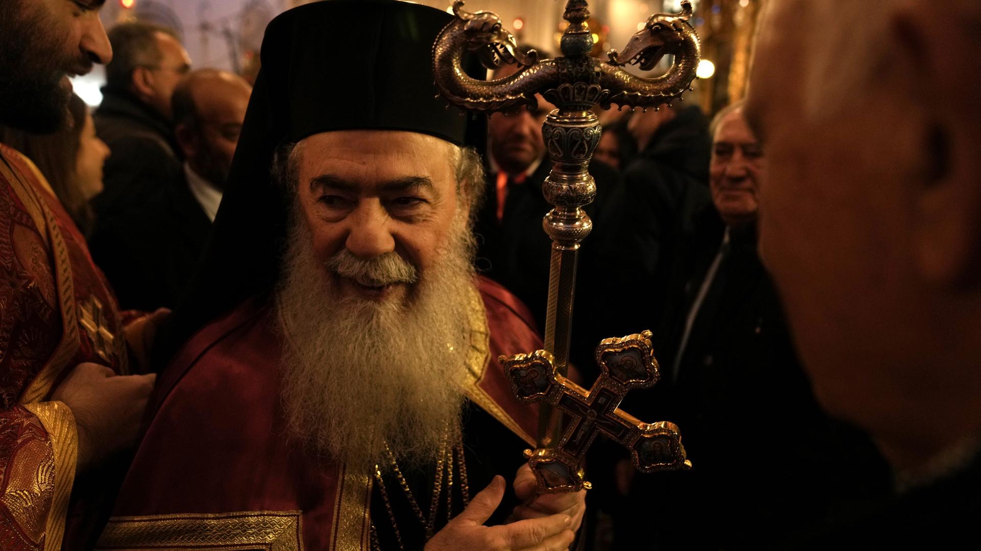 Der griechisch-orthodoxe Patriarch von Jerusalem, Theophilos III. hält ein goldenes Kreuz und seinen Patriarchenstab in der Hand. Er läuft in vollem Ornat durch eine Menge von Gläubigen.