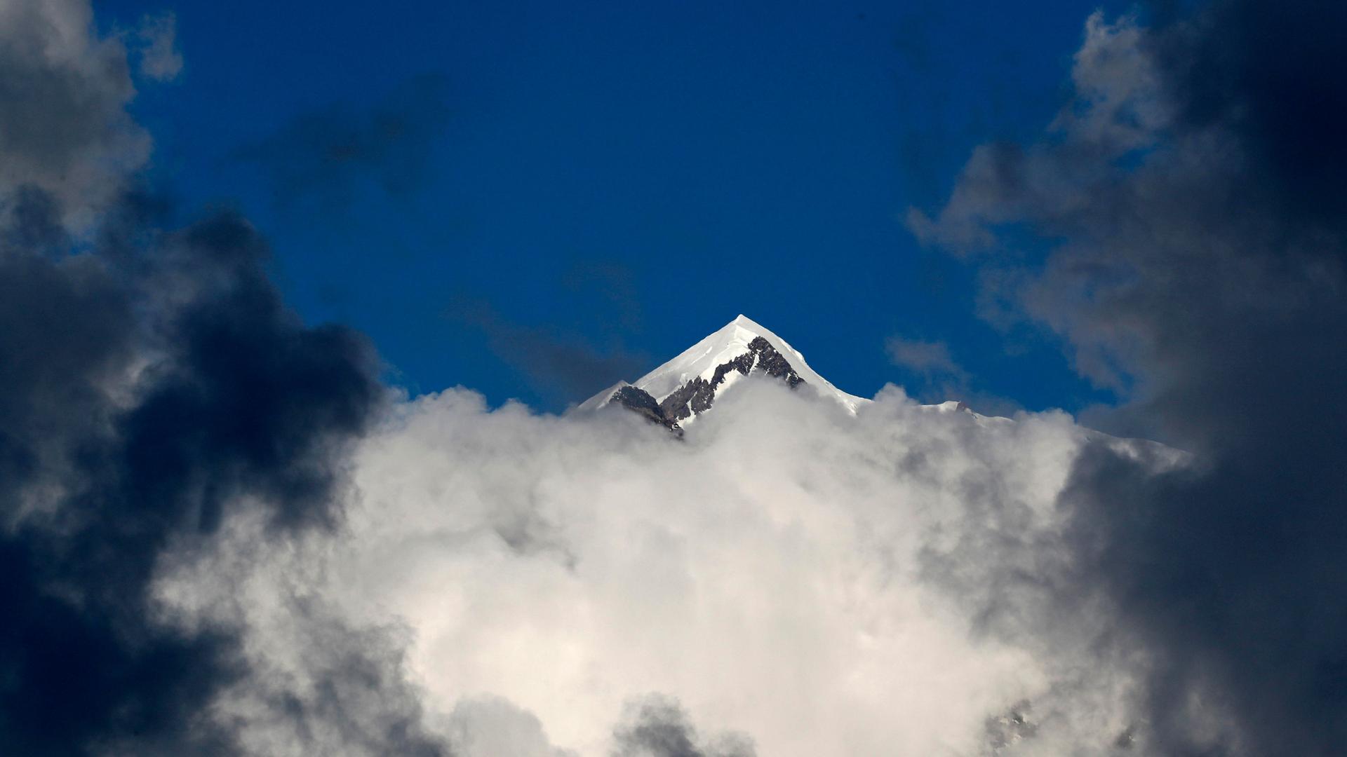 Eine verschneite Bergspitze des Mont-Blanc-Massivs ragt über grau-weiße Wolken in den tiefblauen Himmel empor.