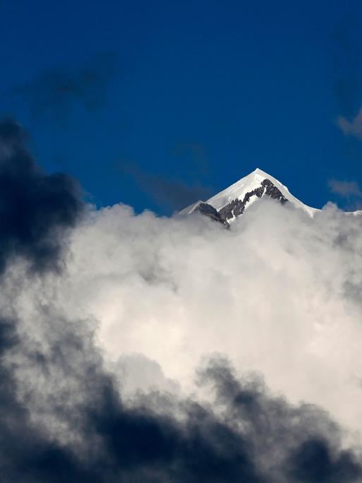 Eine verschneite Bergspitze des Mont-Blanc-Massivs ragt über grau-weiße Wolken in den tiefblauen Himmel empor.