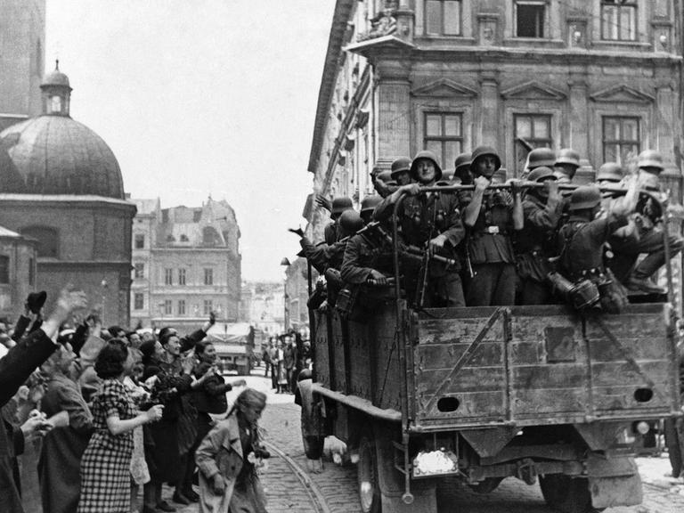 Soldaten der deutschen Wehrmacht fahren am 2. Juli 1941 in einem Militärfahrzeug in die Stadt Lviv, die in der heutigen Ukraine liegt. Am Rand stehen Menschen und jubeln ihnen zu.