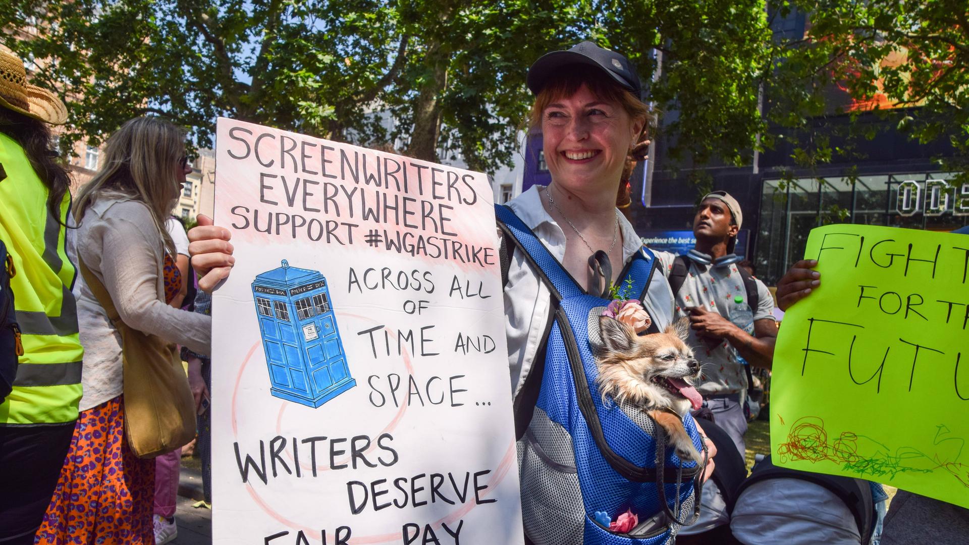 Eine Frau hält ein Transparent hoch, auf dem steht: "Drehbuchautoren in aller Welt, unterstützt den #WGA-Streik. Drehbuchschreiber verdienen eine faire Bezahlung." Im Hintergrund sind weitere Menschen mit Plakaten zu sehen.