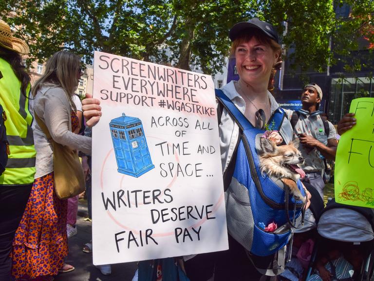 Eine Frau hält ein Transparent hoch, auf dem steht: "Drehbuchautoren in aller Welt, unterstützt den #WGA-Streik. Drehbuchschreiber verdienen eine faire Bezahlung." Im Hintergrund sind weitere Menschen mit Plakaten zu sehen.