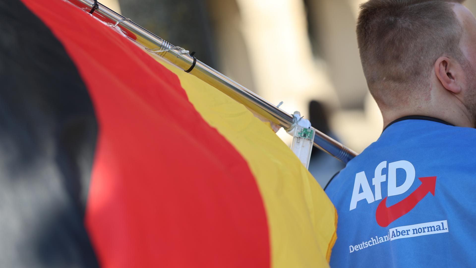 Ein Mann trägt eine Deutschlandfahne und eine Weste, auf der das AfD-Logo und die Aufschrift "Deutschland. Aber normal" zu sehen sind. 