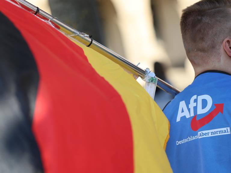 Ein Mann trägt eine Deutschlandfahne und eine Weste, auf der das AfD-Logo und die Aufschrift "Deutschland. Aber normal" zu sehen sind. 