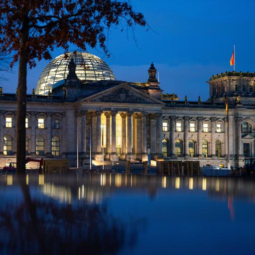 Die Reichstagskuppel in der Abenddämmerung. Es weht eine Deutschlandfahne vor dem blauen Himmel. 