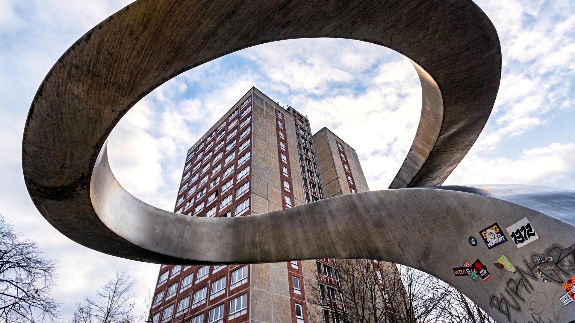 Durch eine geschlungene Skulptur hindurch ist ein Plattenbau zu sehen.