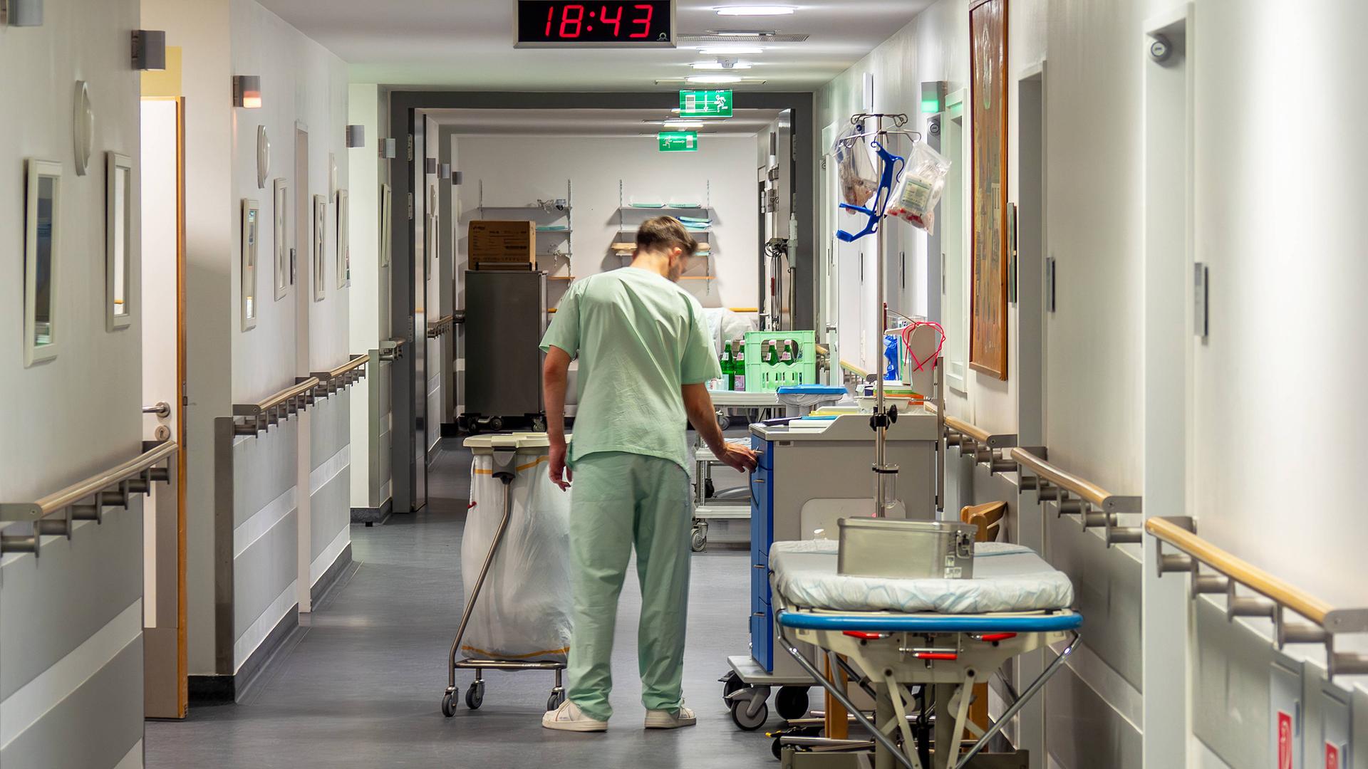 Ein Pfleger in hellgrüner Krankenhauskleidung geht einen Gang entlang und hantiert mit einem großen Abfallbehälter.