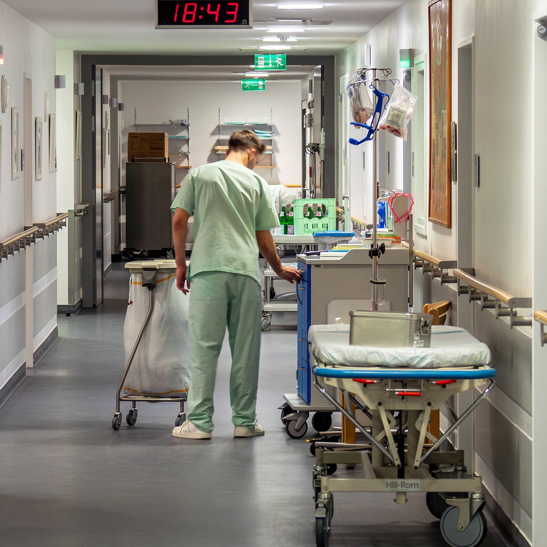 Ein Pfleger in hellgrüner Krankenhauskleidung geht einen Gang entlang und hantiert mit einem großen Abfallbehälter.