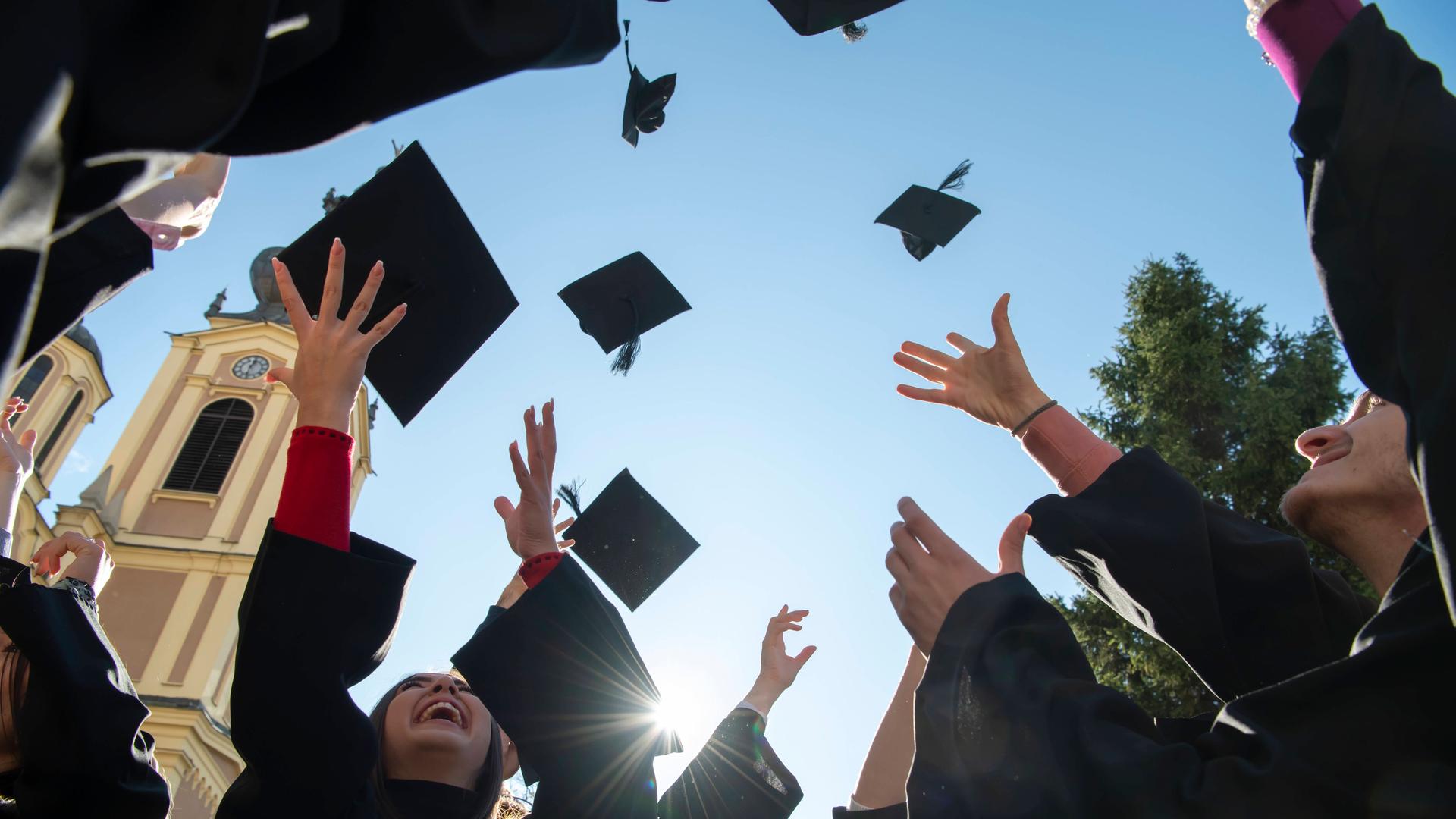 Internationale Studierende in traditionellen schwarzen Roben werfen ihre Absolventenhüte in den blauen Himmel. Symbolbild