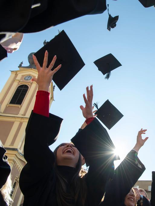 Internationale Studierende in traditionellen schwarzen Roben werfen ihre Absolventenhüte in den blauen Himmel. Symbolbild