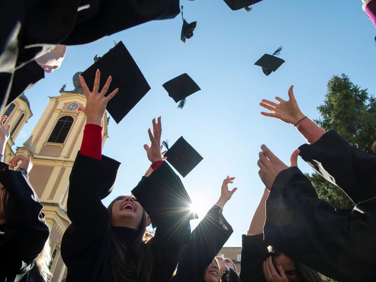 Internationale Studierende in traditionellen schwarzen Roben werfen ihre Absolventenhüte in den blauen Himmel. Symbolbild