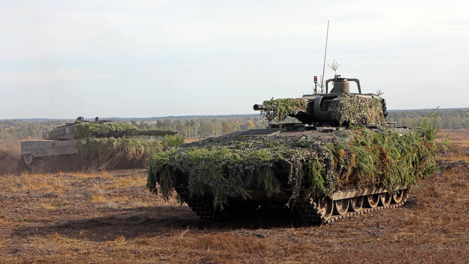 Ein getarnter Schützenpanzer Puma vorne und ein Kampfpanzer Leopard 2 hinten auf einem Truppenübungsplatz bei einer Gefechtsvorführung.