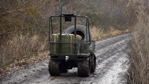 Ein Bodenroboter in Form eines kleinen Wagens auf vier Gummirädern fährt über einen Weg und transportiert einen Reifen und eine Holzkiste. Am Griff des Wagens befindet sich eine Steuerung.