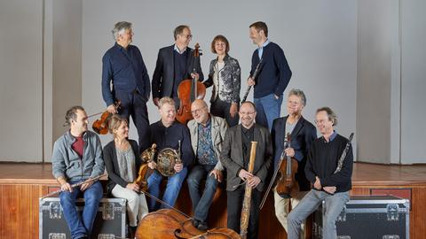 Das Linos Ensemble steht und sitzt mit seinen Instrumenten auf einer Bühne und zum Teil auf Instrumentenkoffern.