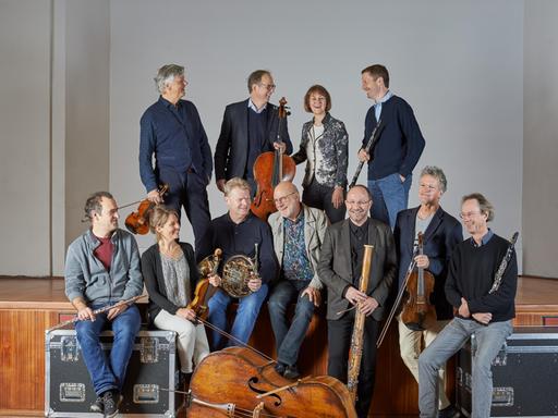 Das Linos Ensemble steht und sitzt mit seinen Instrumenten auf einer Bühne und zum Teil auf Instrumentenkoffern.